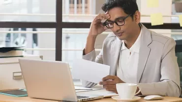 Man in office reading paper with laptop and coffee