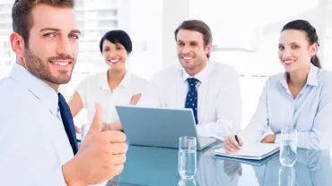 Uśmiechnięty biznesmen pokazujący kciuk w górę z kolegami w tle.