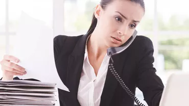 Kobieta wykonuje wiele zadań, rozmawia przez telefon, trzyma papiery, patrzy na laptopa.