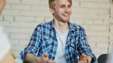 Mężczyzna w koszuli w kratę uśmiecha się podczas rozmowy z innymi.
