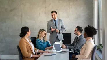 Grupa profesjonalistów na spotkaniu, jedna osoba przemawia.