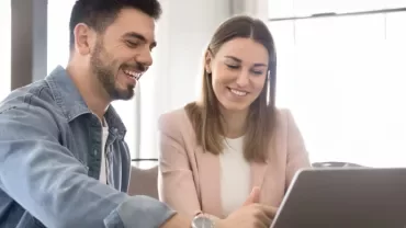 Dwie osoby siedzą przy stole naprzeciw laptopa i uśmiechają się.