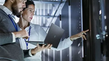 Two people reviewing data on a laptop in a server room.