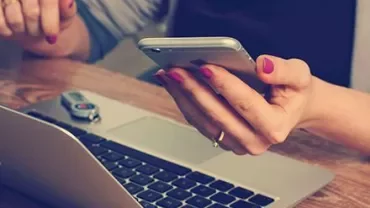 Person holding a smartphone next to a laptop on a wooden desk.