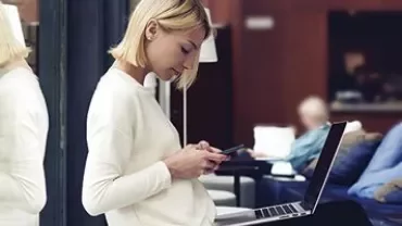 Woman using smartphone with laptop in lap indoors.