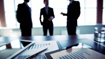 Three businesspeople discuss near a table with charts and tablets.