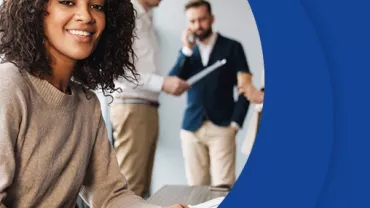 Woman smiling at desk with colleagues in background.