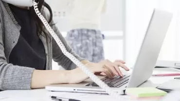 Person typing on laptop while talking on phone at a desk.