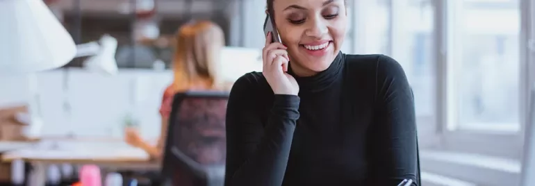 Kobieta w czarnym golfie rozmawia przez telefon w biurze.
