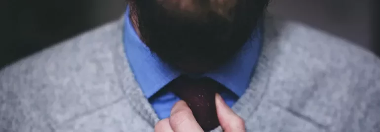 Close-up of a person adjusting a tie, wearing a blue shirt and gray sweater.