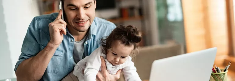 Man multitasking on phone and holding a child near laptop.