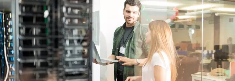 Two people discuss using a laptop near server racks.