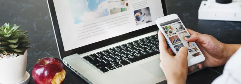 Osoba używająca smartfona i laptopa z nadgryzionym jabłkiem i rośliną na stole.