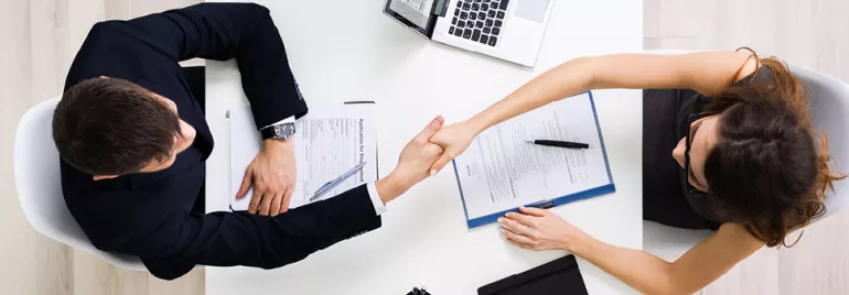 Two people at a desk shaking hands over documents and a laptop.