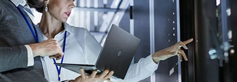 Two people reviewing data on a laptop in a server room.
