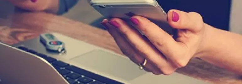 Person holding a smartphone next to a laptop on a wooden desk.