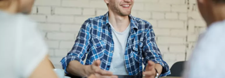 A man in a plaid shirt smiling while sitting at a table with two others.
