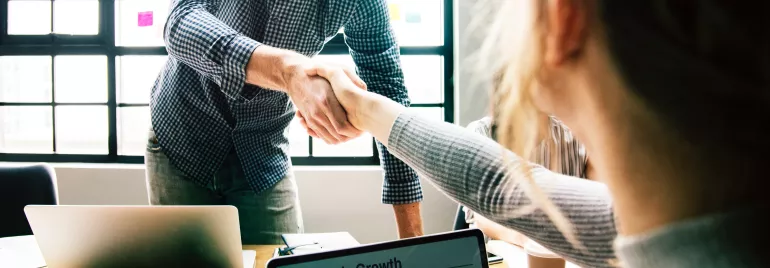 Two people shaking hands in an office with laptops on the table.