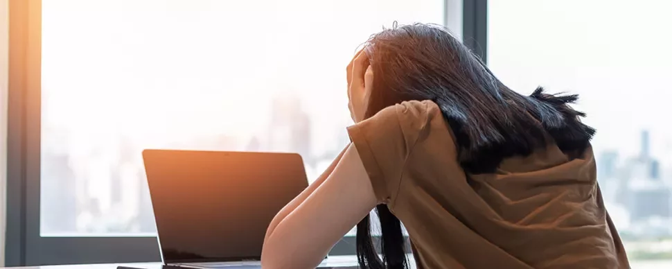 Person stressed at desk with laptop by window.