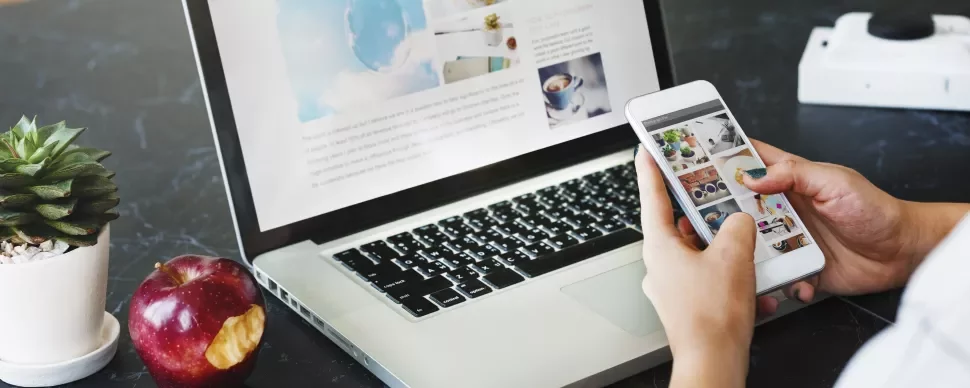 Osoba używająca smartfona i laptopa z nadgryzionym jabłkiem i rośliną na stole.