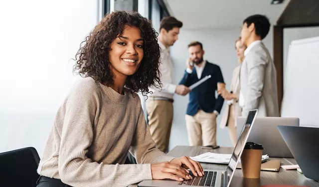Smiling woman working on laptop with colleagues in background.
