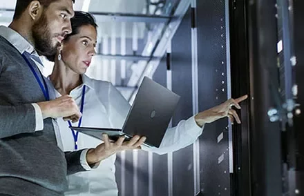 Two people reviewing data on a laptop in a server room.