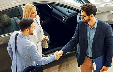 Three people shaking hands near a car in a showroom.
