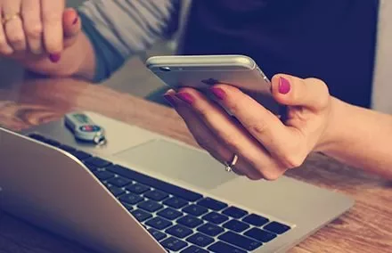 Person holding a smartphone next to a laptop on a wooden desk.